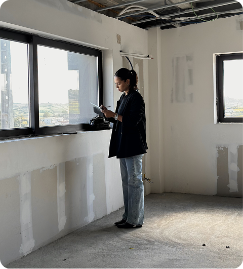 Woman standing in an unfinished interior space near a large window, writing notes on a clipboard.